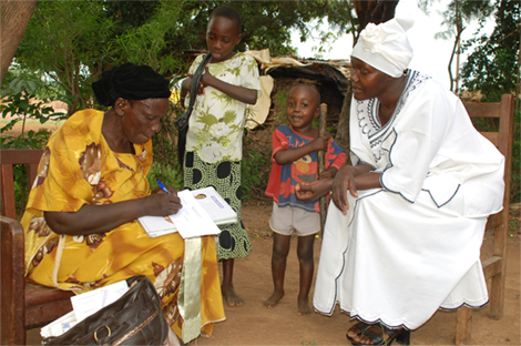 Photo of a female health worker talking to a seated woman with two children