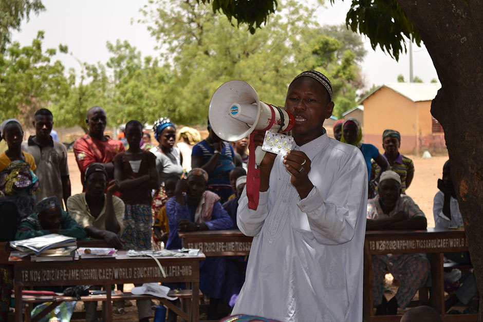 Photo of a man with a bullhorn, holding a pack of pills, under a tree with villagers in the background.