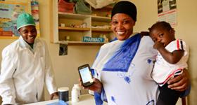 Photo of a woman in a health clinic holding a smartphone and an infant.